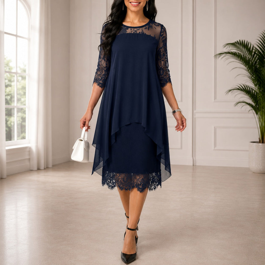 Femme portant la robe bleu marine en mousseline superposée, manches trois-quarts en dentelle et ourlet dentelle, avec sac blanc, dans une pièce lumineuse. 1