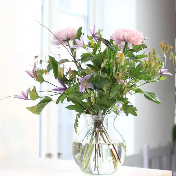 Bouquet aux fleurs roses et violettes dans un vase rond transparent, tiges entrecroisées visibles dans l'eau près d'une fenêtre. 4