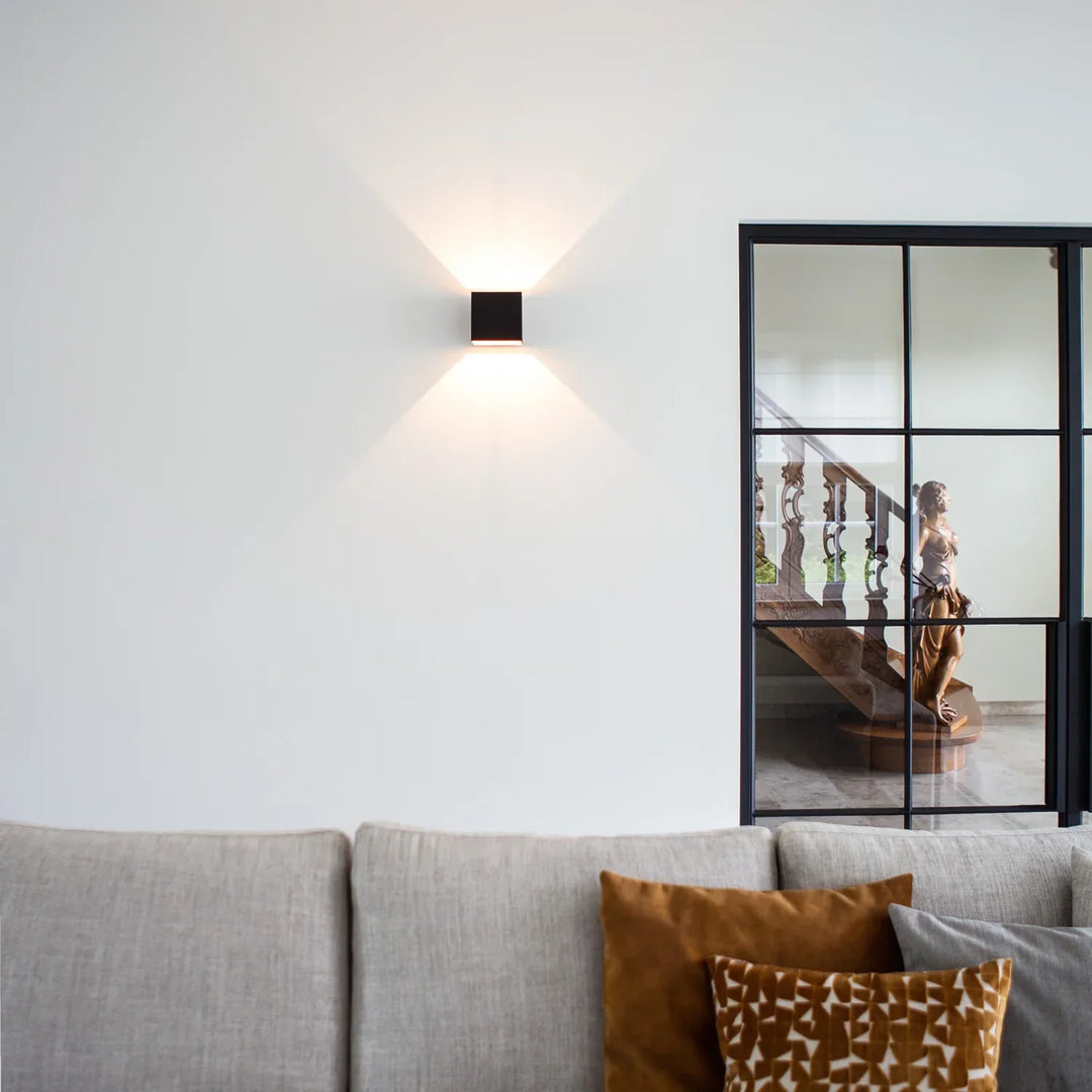Applique cubique noire allumée sur un mur blanc au-dessus d'un canapé gris à coussins ocre, près d'une porte vitrée à verrière. 10
