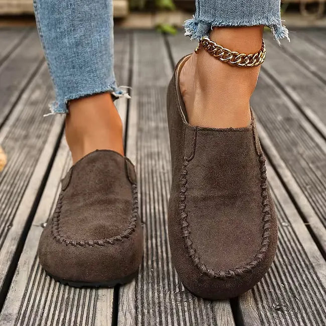 Vue de face de mules marron foncé façon daim portées avec un jean effiloché et une chaînette dorée à la cheville. 4