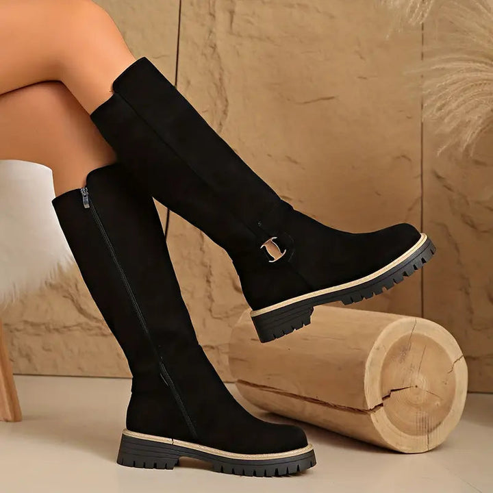 Bottes noires hautes portées jambes croisées, vue de profil montrant la fermeture éclair sur le côté et la semelle crantée. 2