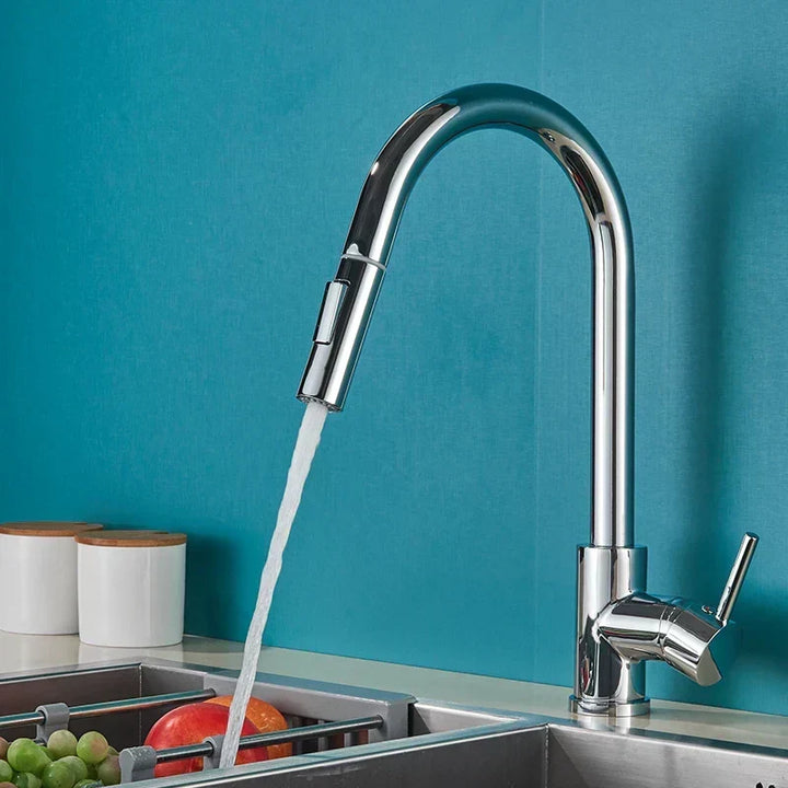 Robinet chromé à bec haut et douchette, jet d'eau coulant vers un évier avec des fruits, devant un mur bleu-vert. 1