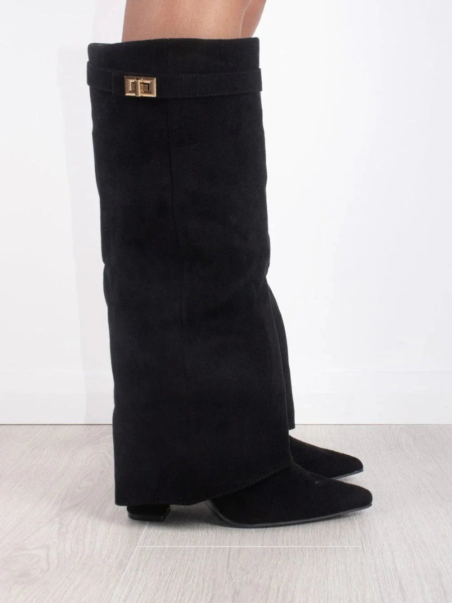 Vue de profil d'une botte haute noire en daim avec languette repliée et boucle dorée, portée sur un parquet clair. 1