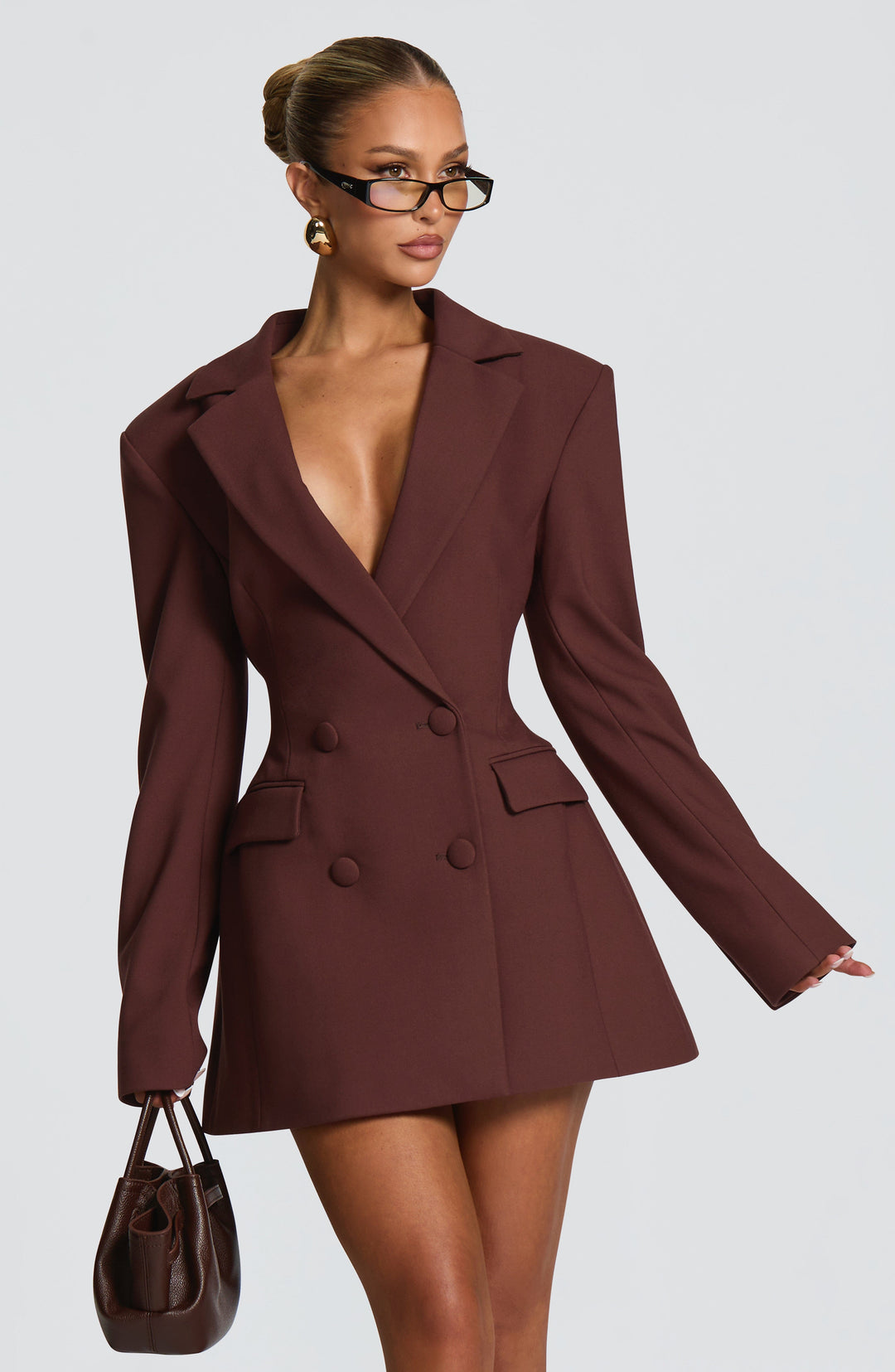 Robe blazer marron portée en trois quarts par une mannequin tenant un petit sac en cuir marron, silhouette cintrée et jupe évasée. 2