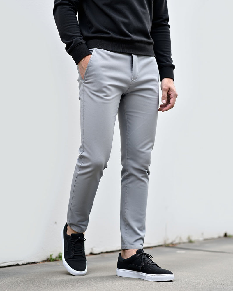 Chino gris clair porté devant un mur blanc avec un pull noir et des baskets noires à semelle blanche. 2