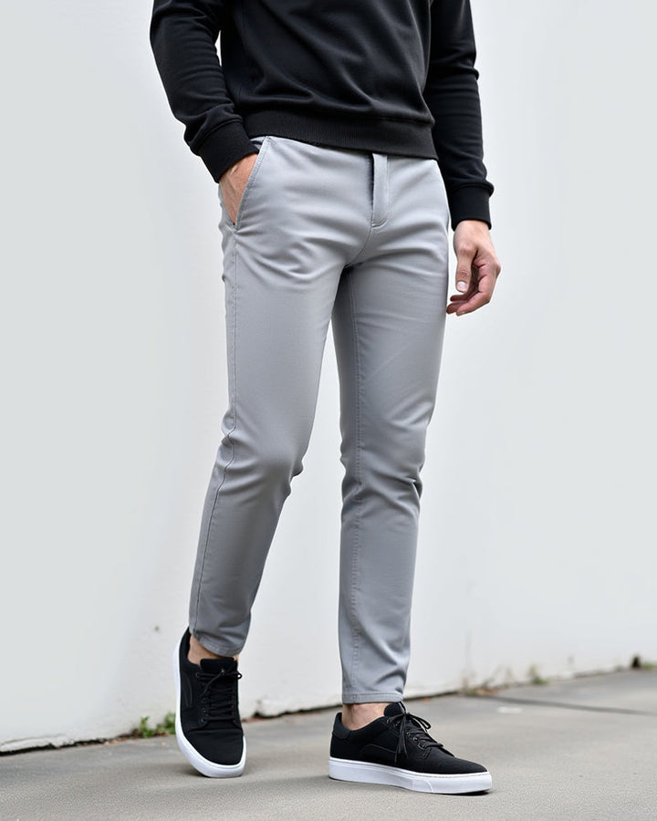 Chino gris clair porté devant un mur blanc avec un pull noir et des baskets noires à semelle blanche. 2