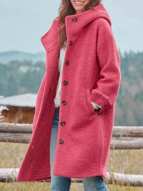Femme souriante portant le manteau long à capuche rose corail, boutonné devant, une main dans la poche, devant une clôture en bois. 1