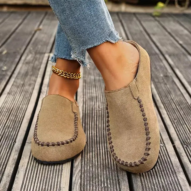 Vue de face de mules beige façon daim à surpiqûres brunes, portées en marchant sur une terrasse en bois avec un jean. 5