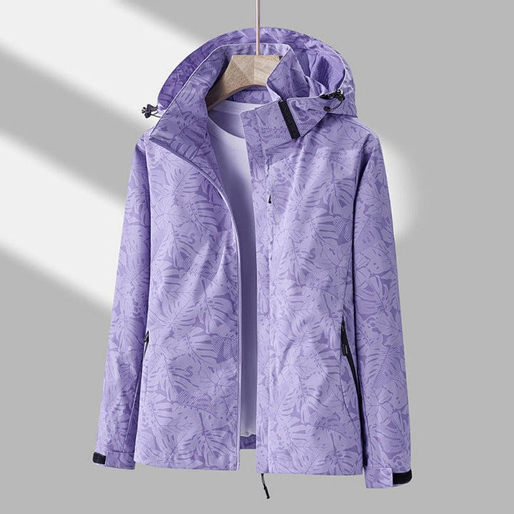 Veste violet lilas à motif de feuilles tropicales, capuche relevée, zip ouvert sur un t-shirt clair, présentée sur cintre en bois. 1