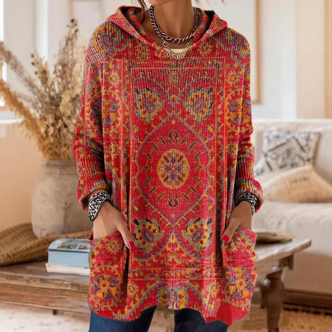 Tunique rouge à capuche en maille côtelée, motif de tapis oriental jaune et bleu, portée mains dans les poches dans un salon. 1