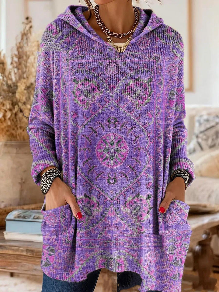 Version violette de la tunique à capuche, motif de tapis rose et vert sur maille côtelée, portée avec les mains dans les poches. 2