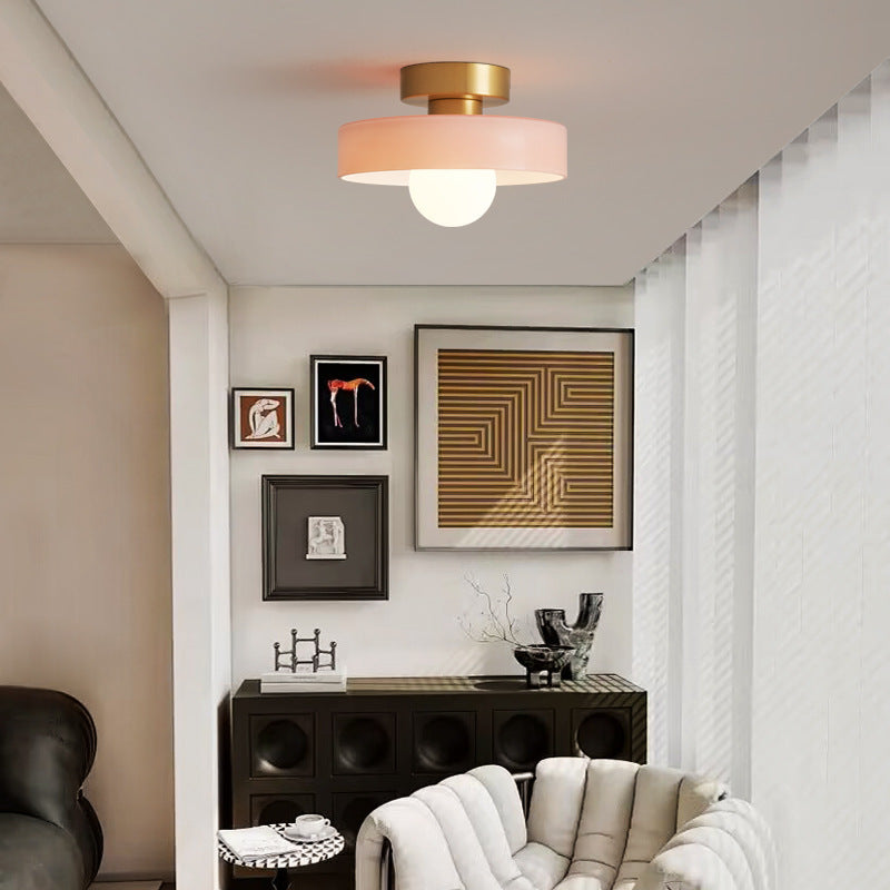 Plafonnier à abat-jour rose et base dorée, allumé dans un salon décoré de tableaux, avec un buffet noir et un fauteuil bouclé. 3