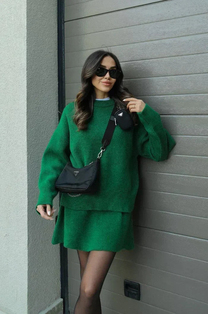 Mannequin en ensemble vert, adossée au portail gris, une main relevée vers le visage, sac bandoulière noir porté en travers. 10