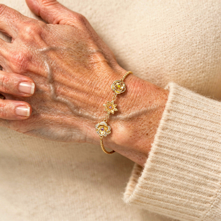 Main et poignet d'une femme âgée portant le bracelet doré aux trois fleurs, manche de pull beige en maille douce. 11