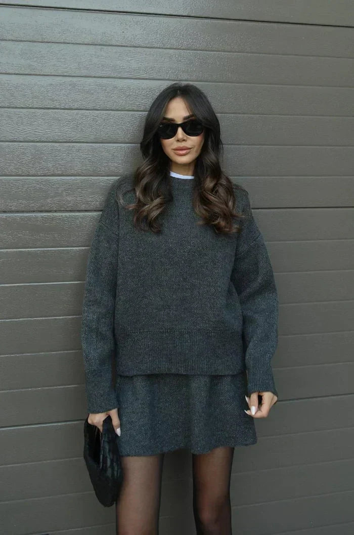 Ensemble gris anthracite en maille porté de face contre un mur gris, avec lunettes noires et pochette noire à la main. 11