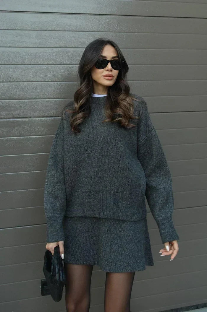 Pull ample et jupe courte évasée gris anthracite, cheveux ondulés, collants noirs et pochette noire tenue à la main. 12