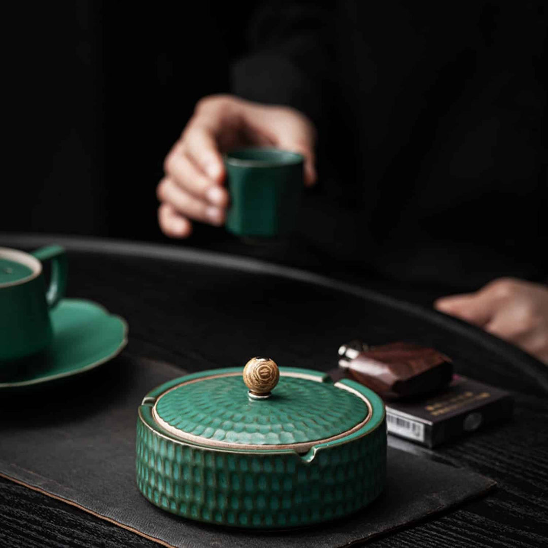 Cendrier vert fermé sur un set de table gris, avec une tasse et une soucoupe vertes et une personne tenant une petite tasse. 4