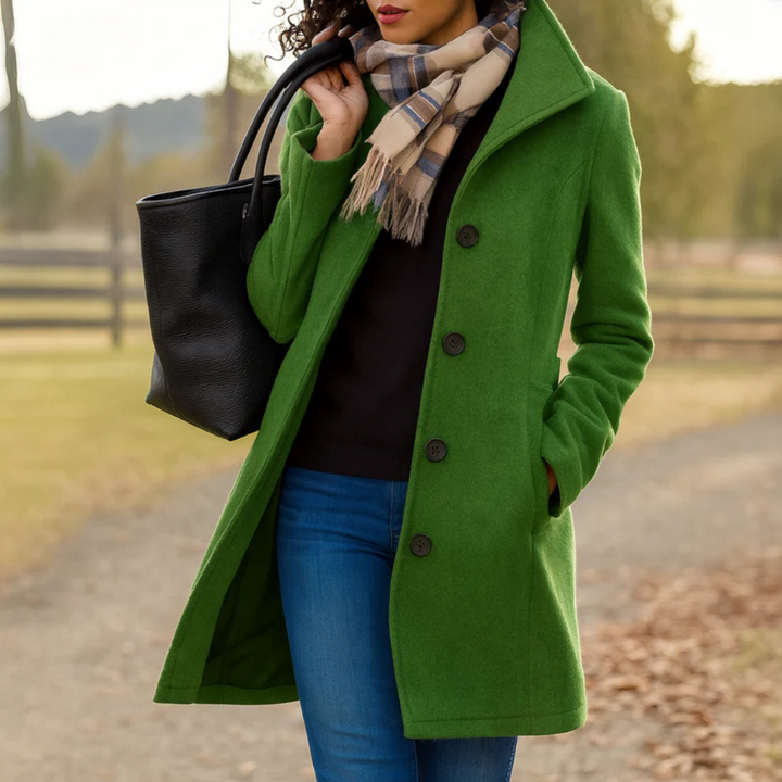 Manteau vert prairie porté par une femme, main dans la poche, avec écharpe à carreaux beige et bleu et sac noir sur un chemin. 6