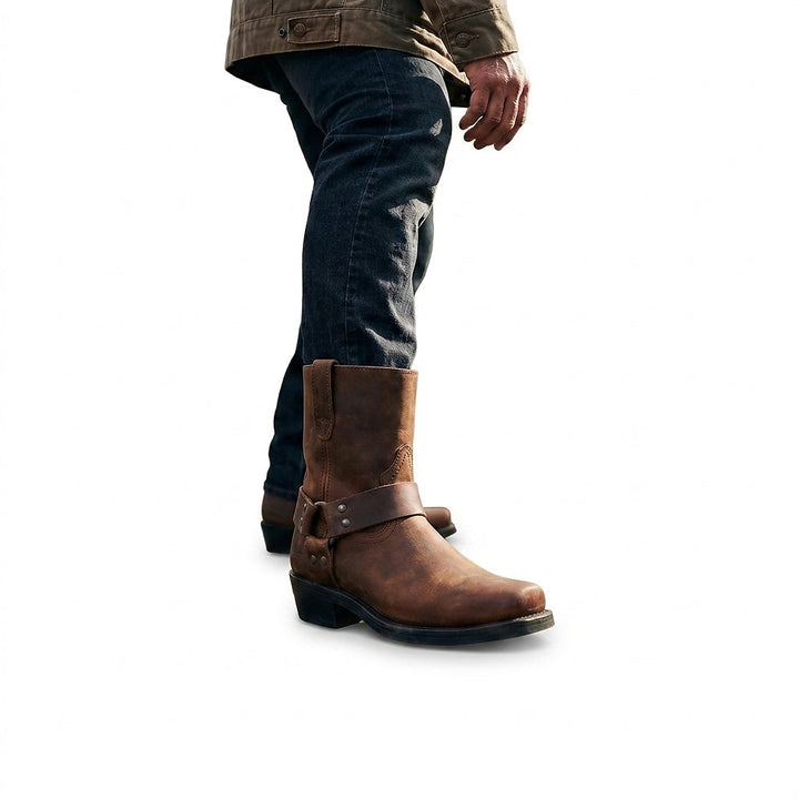 Homme en jean foncé et veste kaki portant la botte marron à sangle cloutée, vu de profil en marchant. 1