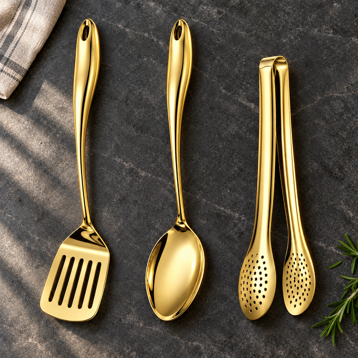 Spatule ajourée, cuillère et pince dorées à manche courbé posées sur une pierre sombre, avec torchon rayé et brin de romarin. 23