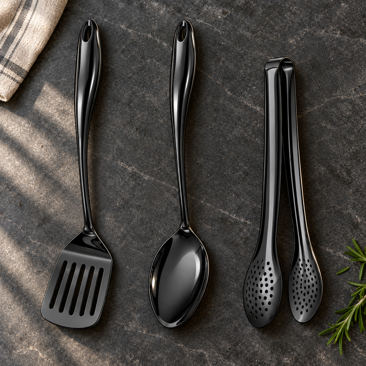 Spatule ajourée, cuillère et pince noires à manche courbé posées sur une pierre sombre, avec torchon rayé et brin de romarin. 27