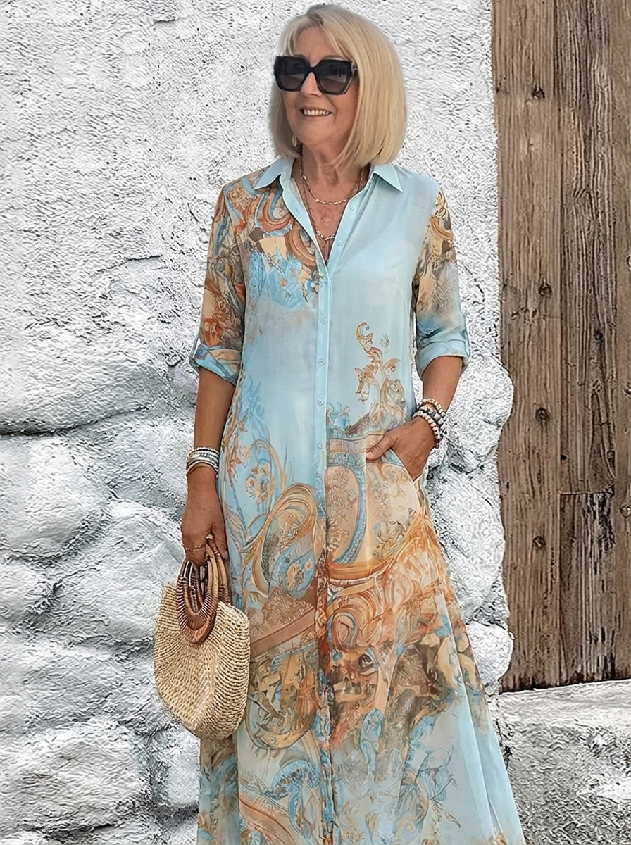Femme en robe chemise longue bleu ciel à motifs baroques orangés, manches retroussées, sac en paille, devant un mur blanc. 1