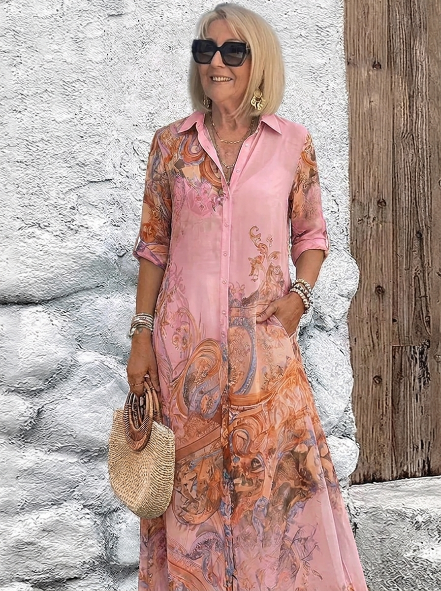 Robe chemise longue portée en rose à motifs baroques orangés, mannequin souriante, lunettes de soleil, une main dans la poche. 4