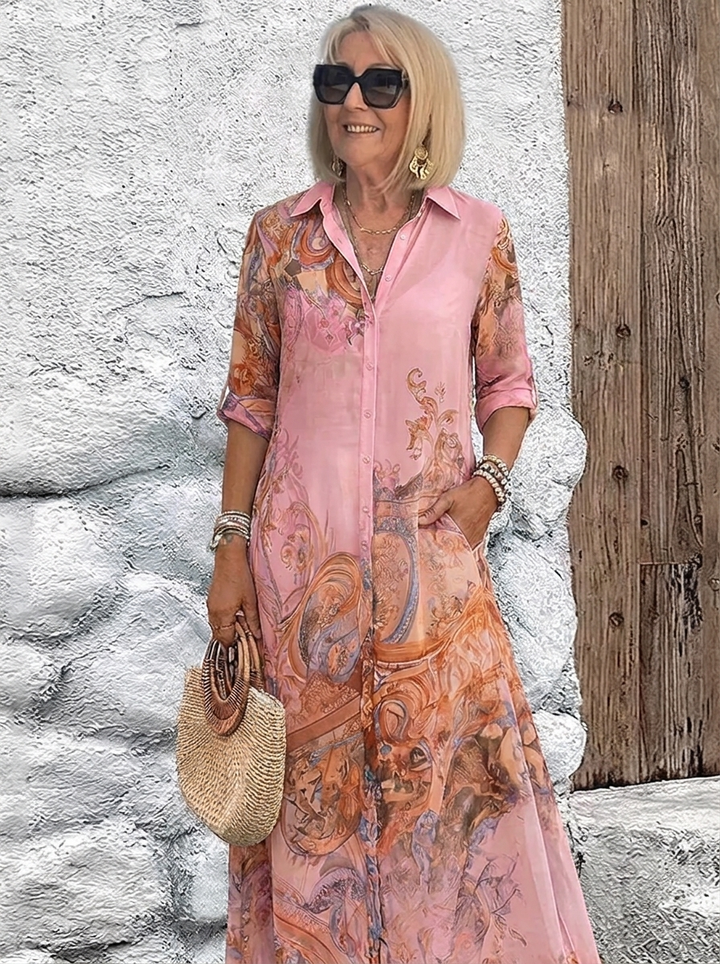 Robe chemise longue portée en rose à motifs baroques orangés, mannequin souriante, lunettes de soleil, une main dans la poche. 4