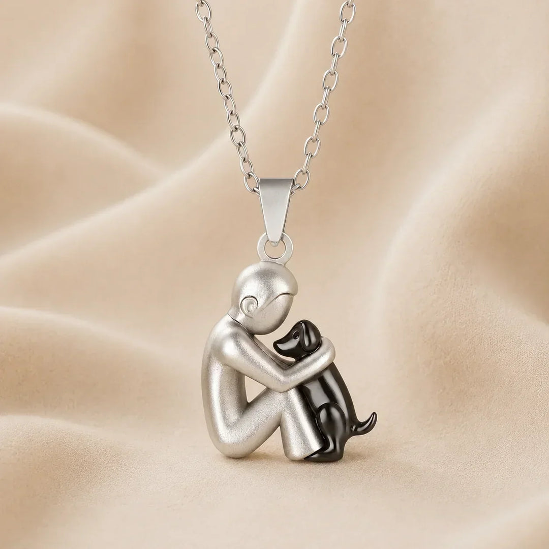 Pendentif mat d'une personne argentée recroquevillée serrant un petit chien noir dans ses bras, photographié sur un tissu beige. 7