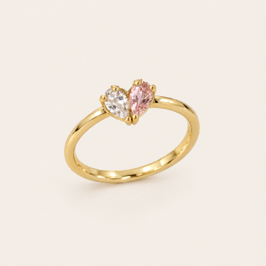 Bague dorée vue de trois quarts, avec une pierre transparente et une pierre rose en forme de goutte qui dessinent un cœur. 1