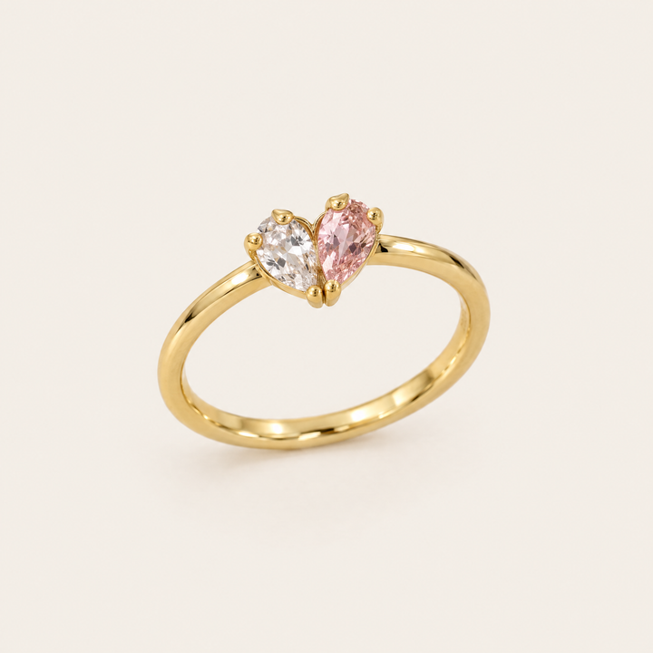 Bague dorée vue de trois quarts, avec une pierre transparente et une pierre rose en forme de goutte qui dessinent un cœur. 1