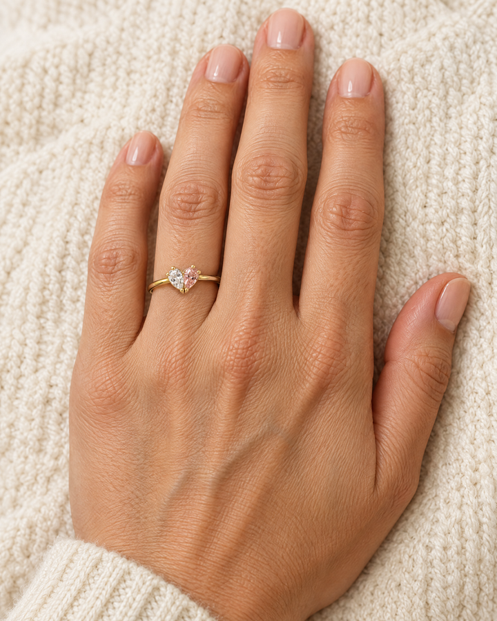 Bague dorée portée à l'annulaire d'une main posée sur un tricot écru, avec son cœur de pierres transparente et rose. 2