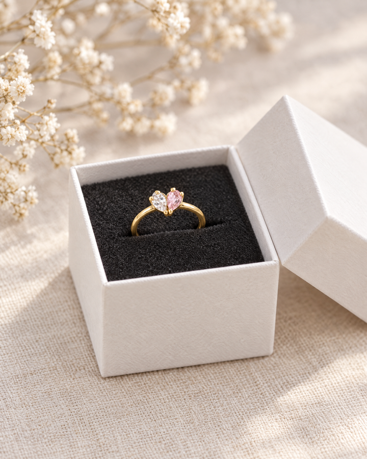 Bague dorée aux pierres transparente et rose dans un écrin blanc ouvert à l'intérieur noir, près de fleurs séchées. 3
