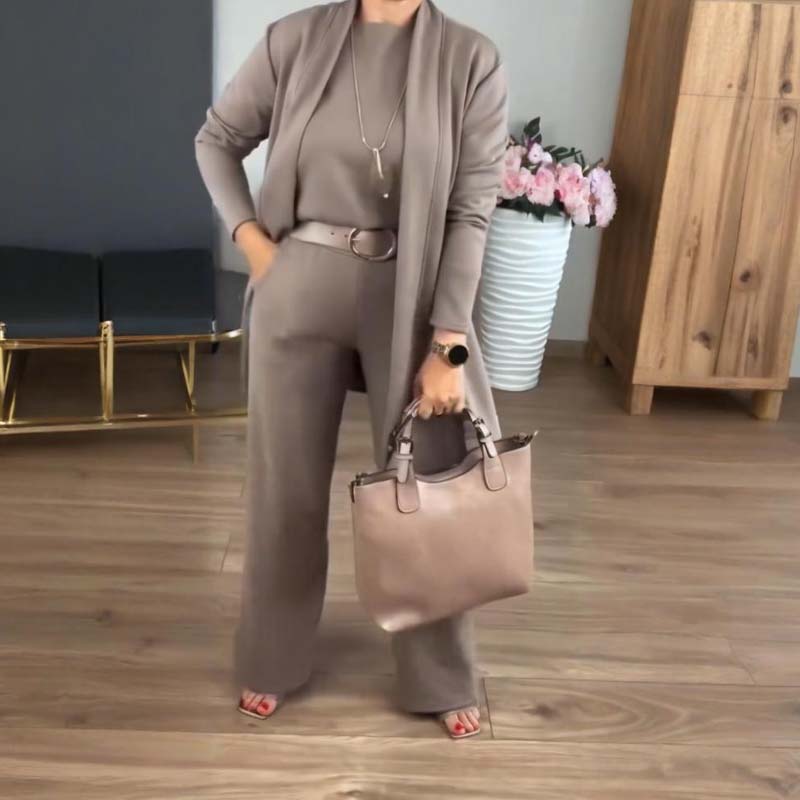 Femme portant l'ensemble taupe avec ceinture et long collier, tenant un sac beige, devant un vase de fleurs roses. 10