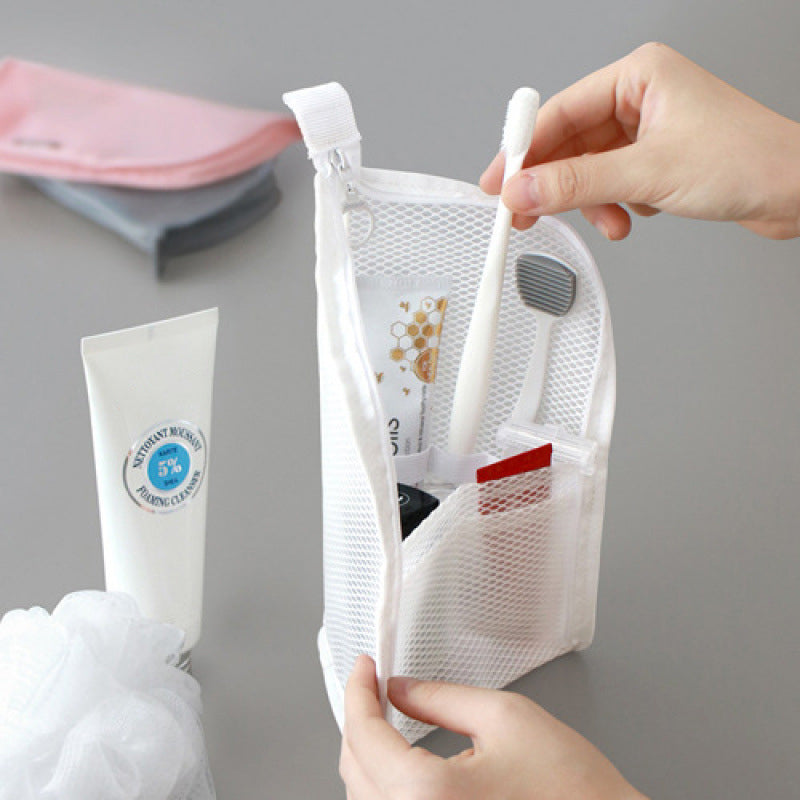 Mains sortant une brosse à dents de la pochette blanche en filet ouverte, avec tube et accessoires de toilette posés autour. 1