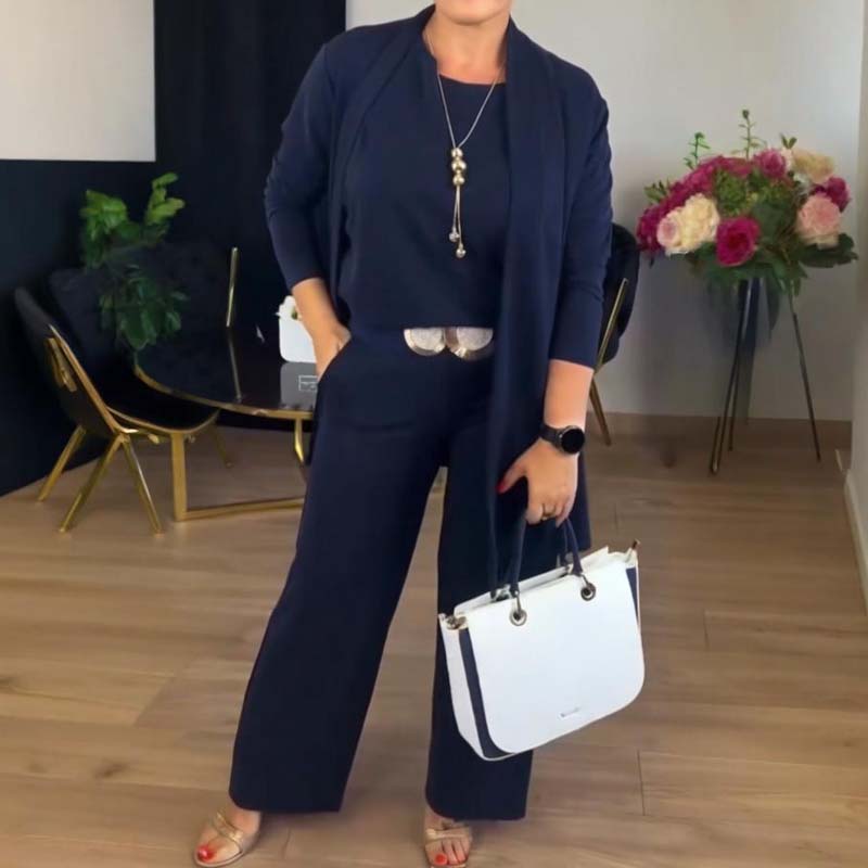 Femme en ensemble bleu marine avec long collier doré et ceinture dorée, tenant un sac blanc, dans un intérieur décoré de fleurs. 9
