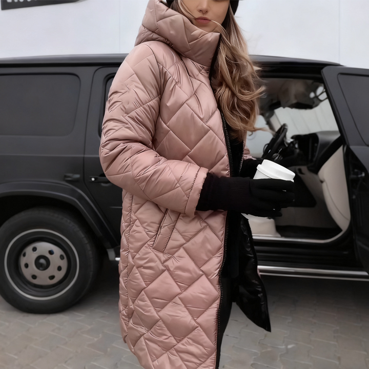 Femme portant le manteau matelassé à capuche rose poudré, motif losanges, zip ouvert, gants noirs et gobelet à la main devant un véhicule noir. 1
