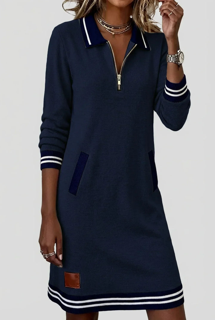 Robe bleu marine portée par un mannequin, main levée vers le visage, zip doré au col et poches contrastées. 9