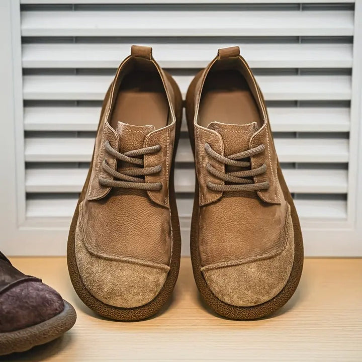 Paire de chaussures à lacets en cuir velours camel, vue de face, posée devant un volet blanc. 1
