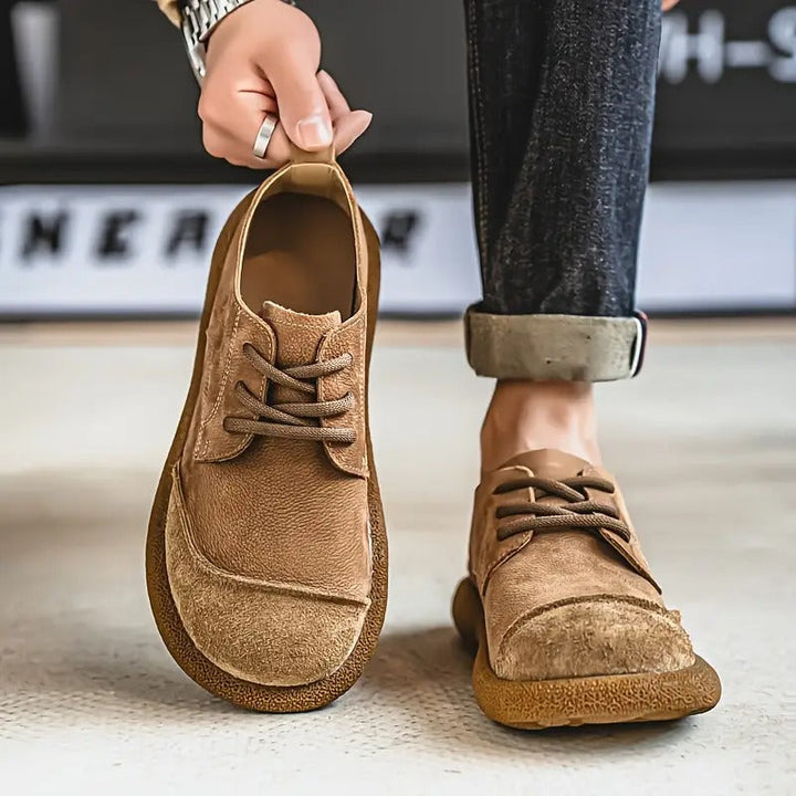 Homme tenant une chaussure camel par la languette arrière, l'autre chaussure au pied, jean retroussé. 3