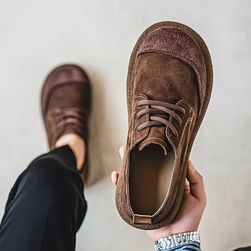 Chaussure marron tenue en main vue d'en haut, laissant voir l'intérieur et les coutures claires, l'autre pied chaussé en arrière-plan. 7