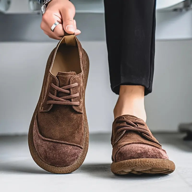 Modèle marron foncé : une chaussure tenue par la languette, l'autre portée avec un pantalon noir, bout arrondi en daim. 6