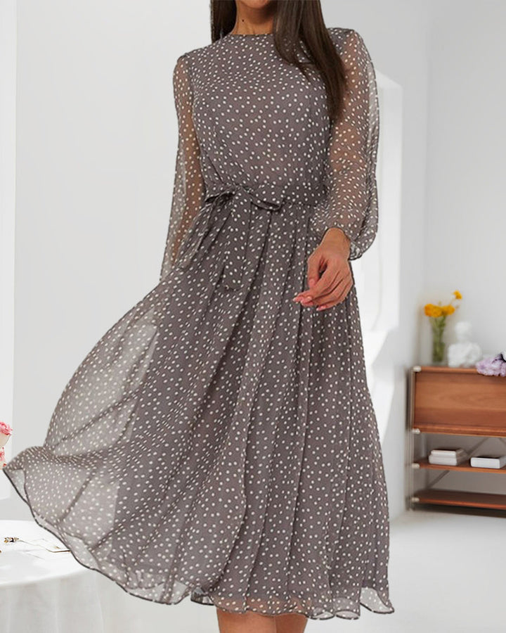 Mannequin en robe grise à pois et ceinture nouée dans un intérieur lumineux, avec étagère en bois et fleurs jaunes. 3