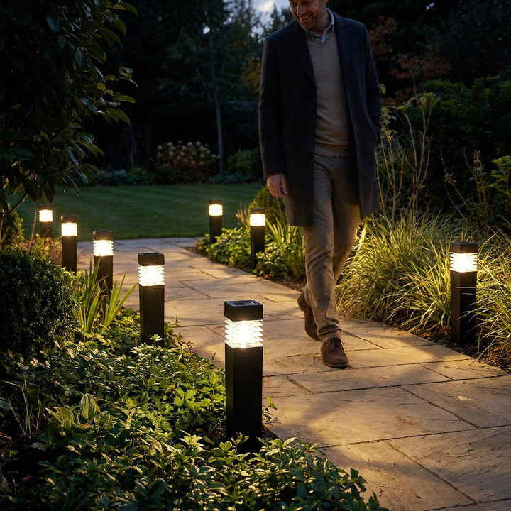 Rangée de bornes lumineuses noires allumées le long d'une allée dallée de jardin la nuit, avec un homme en manteau qui marche. 1
