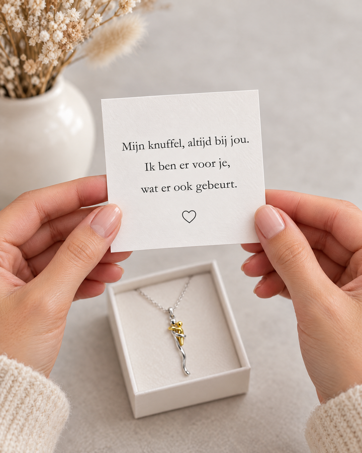 Deux mains tiennent une carte de message au-dessus de l'écrin ouvert contenant le collier, près d'un vase de fleurs séchées. 6