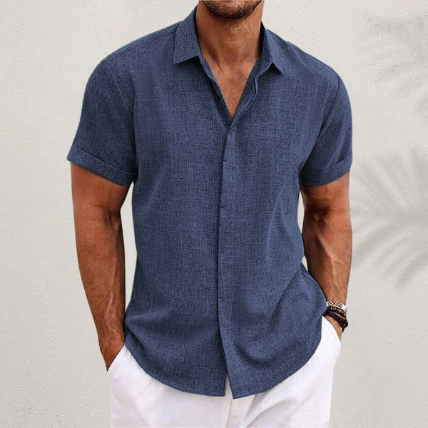 Chemise bleu marine à manches courtes retroussées, portée par un homme en pantalon blanc devant un mur clair. 6