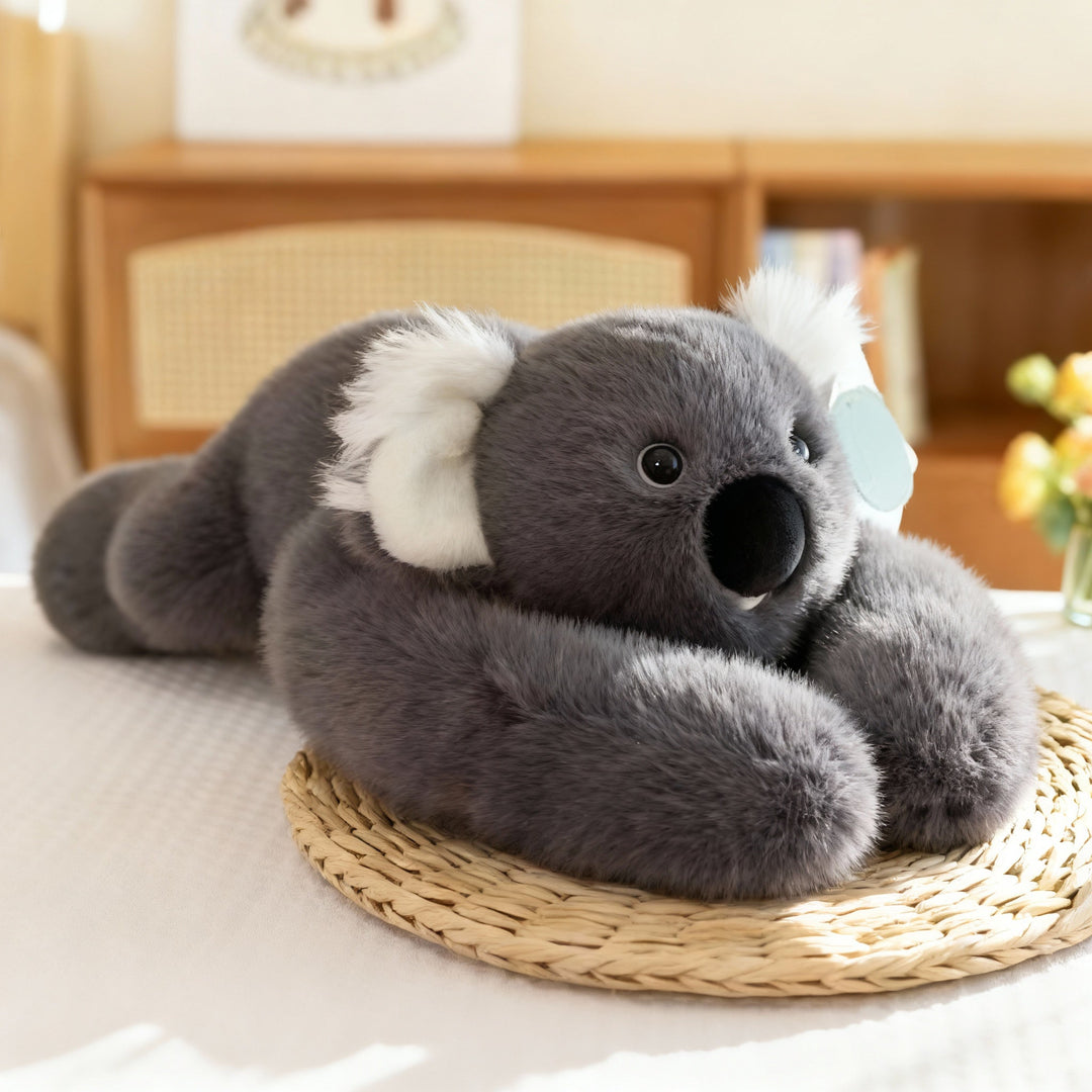 Koala en peluche gris foncé aux grandes oreilles blanches, allongé sur un set de table en paille, longs bras repliés devant lui. 16
