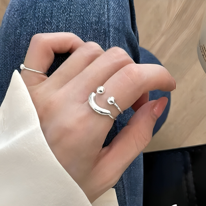 Bague argentée ouverte en forme de visage souriant portée à l'index, avec une chemise blanche et un jean en arrière-plan. 1