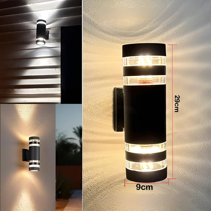Applique cylindrique noire à bandes transparentes, hauteur 29 cm et diamètre 9 cm, montrée allumée avec deux vues d'ambiance sur des murs extérieurs. 4