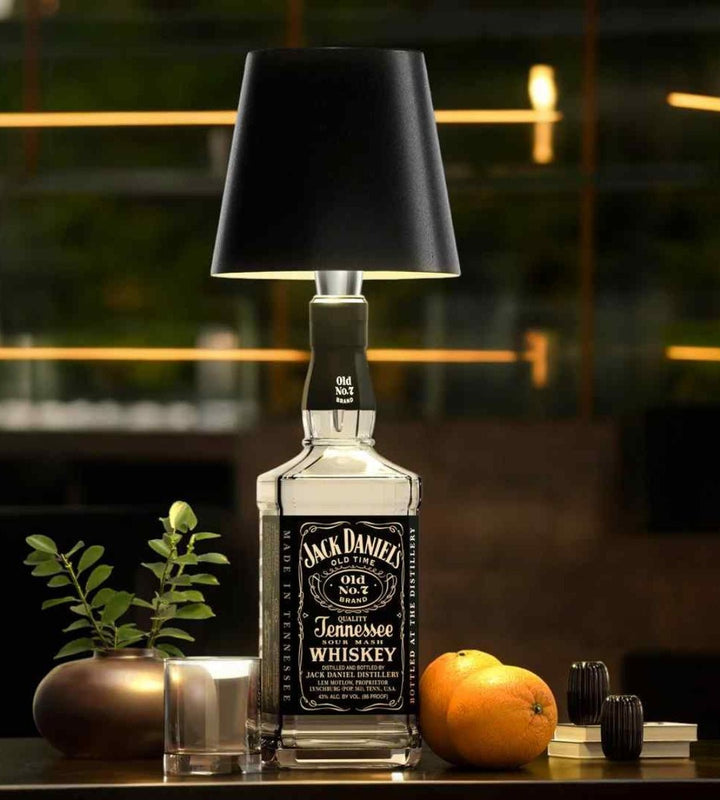 Abat-jour noir fixé sur une bouteille de whisky carrée, posée sur un comptoir avec orange, verre et plante. 2